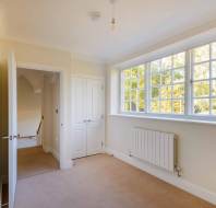 Empty room with beige carpet, white walls, a radiator, and a large window. Door to the left is open, leading to a staircase. Hanging lightbulb without fixture on the ceiling.