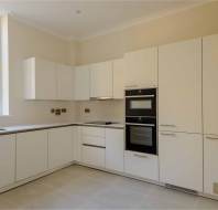 A modern kitchen with white cabinets, a built-in oven, and a window on the left. The floor is tiled, and the walls are painted beige.