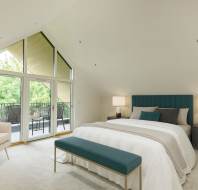 A modern bedroom with a large window and balcony. It includes a bed with a teal headboard, two nightstands with lamps, a beige armchair, and a teal bench at the foot of the bed.