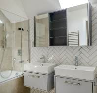 Modern bathroom with herringbone tile wall, dual sinks with storage drawers, mirrored cabinets, and a glass-enclosed bathtub with a rain showerhead.
