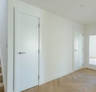 A bright hallway features a white door and a staircase on the left, a second door straight ahead, and a pair of doors with glass panels on the right leading to another room.