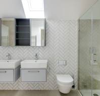 Modern bathroom with herringbone tile, dual sinks, a toilet, and a glass shower. Skylight above and a mirrored cabinet on the wall.