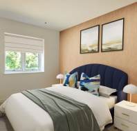 A modern bedroom with a bed featuring blue pillows, a green throw, and a padded headboard. Two framed artworks hang above, with bedside lamps on white nightstands. A window and plant are on the left.