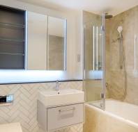 Modern bathroom with a wall-mounted sink, illuminated mirror, tiled walls, bathtub, and glass shower screen.