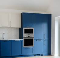 Modern kitchen with blue cabinets, white upper cupboards, a built-in stainless steel oven, and marble backsplash. Light wood flooring and a window are visible.