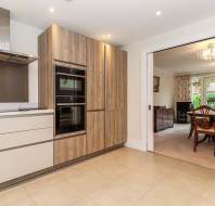 Contemporary kitchen with wood-paneled oven wall, white cabinets, and tile flooring. Adjacent dining room with wooden table and chairs, separated by a sliding door. Chandelier lighting visible.