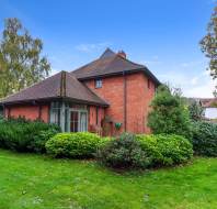 A red brick house with a pitched roof is surrounded by greenery, including shrubs and trees, on a well-maintained lawn under a clear blue sky.