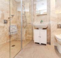 Modern bathroom with a walk-in shower, beige tile walls, white sink on a cabinet, and a toilet. Foldable seat installed in the shower.