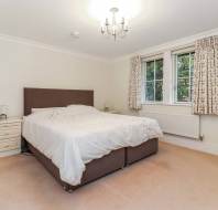 A bedroom with a double bed, white bedding, two bedside tables with lamps, a chandelier, and curtained windows. A wall-mounted mirror is on the right.