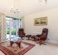 Living room with two brown recliners, an ottoman, a glass-top coffee table on a patterned rug, and a painting on the wall. Glass doors open to an outdoor view.
