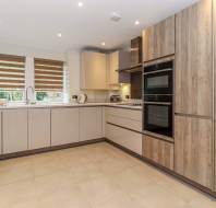 Modern kitchen with beige cabinets, wood-paneled appliances, double oven, and a window with striped blinds. Neutral tiled flooring, small counter appliances, and recessed lighting.