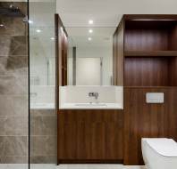 Modern bathroom with a glass shower, beige tiles, and wooden cabinetry. Includes a sink and toilet, all in a minimalist design.