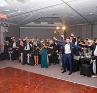 A group of people in formal attire dance with their hands up in a banquet hall with round tables decorated in black.