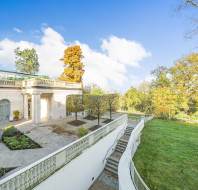 Classical mansion with arched windows, surrounded by trees and a large lawn. Stone staircase leads down from a landscaped terrace. Clear blue sky in the background.
