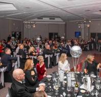 A formal event with people seated at round tables, elegantly dressed in evening attire, with disco balls on the tables and a dance floor in the center.