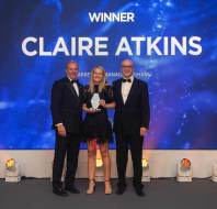 Three people in formal attire stand on a stage. A woman in the middle holds a trophy. A screen behind them displays "Winner Claire Atkins.