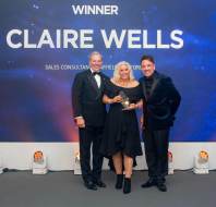Three people stand on stage, one woman holds an award. The backdrop reads "Winner Claire Wells Sales Consultant." They are dressed in formal attire.