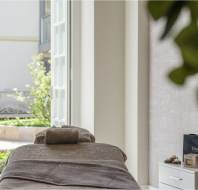 A spa room with a massage table, neatly arranged towels, and a window view of a garden sculpture and greenery.