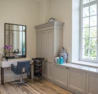 A salon interior with two styling stations, mirrors, chairs, and a storage cabinet against a wall. A window on the right lets in natural light.
