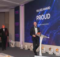 Two men in tuxedos on stage during an awards ceremony. One man is speaking at a podium with "Values Award Proud" displayed on a screen in the background.