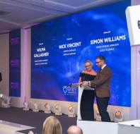 Two people stand at a podium on stage during an awards event. A large screen behind them displays the names of finalists. An audience member applauds.