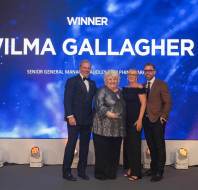 Four people stand on a stage celebrating a winner, with a large background screen displaying the name "Wilma Gallagher.