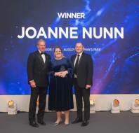 Three people standing on stage under a "Winner Joanne Nunn" sign at an award ceremony.