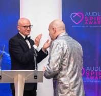 Two men on stage at the Audley Spirit Awards; one in a tuxedo claps, the other in a shiny silver jacket.