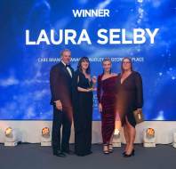 Four people pose on stage with a trophy in front of a large screen displaying "Winner Laura Selby.