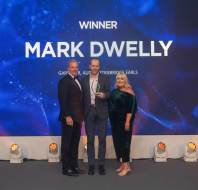 Three people stand on stage with one holding an award. A large display in the background reads "Winner Mark Dwelly.