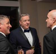 Three men in formal attire holding glasses of champagne engage in conversation at an indoor event.