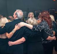 A group of people at a party embrace with their backs to the camera, while others are visible in the background.