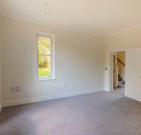 An empty room with beige walls, a gray carpet, a window showing greenery outside, a radiator beneath it, and an open door leading to a hallway with stairs.