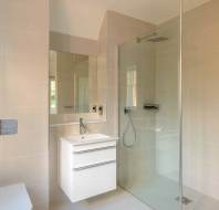 Minimalist bathroom with beige tiles, featuring a glass-enclosed shower, white vanity with a sink, wall-mounted faucet, mirror, and partial view of a toilet.
