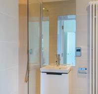 Modern bathroom with glass shower, rectangular mirror, and a white sink with chrome faucet. Smooth beige wall tiles and a wall-mounted toilet paper holder are visible.