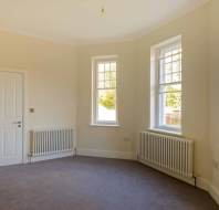 An empty room with light-colored walls, two large windows, a closed white door, and a carpeted floor. Radiators are placed below the windows.