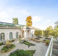 Elegant white building with arched windows, surrounded by a courtyard featuring neatly trimmed hedges, benches, and autumn trees under a clear blue sky.