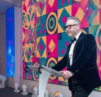 A man in a tuxedo stands at a podium with microphones in front of a colorful geometric background.
