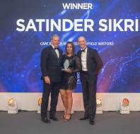 Three people are standing together on a stage. The person in the center is holding an award, with a backdrop displaying "Winner Satinder Sikri.