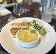 A small dish of pâté with two slices of toasted bread and greens on a white plate, placed on a dark wooden table with glasses and utensils nearby.