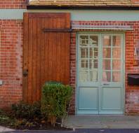 A brick house facade with a pale blue door, wooden sliding barn door, and two windows. Green shrubs are on the left.