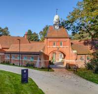 Historic brick building with a clock tower, arched passage, and tile roof, surrounded by trees. A sign is visible on the front lawn.