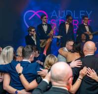 A band in suits performs on stage while a crowd dressed in formal attire dances and embraces each other in front. A bright screen with "Audley Spirit Awards" is in the background.