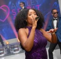 A woman sings into a microphone on stage, with two musicians in the background.