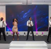 Five musicians performing on stage with microphones and instruments, dressed in formal attire, in front of a large screen displaying abstract patterns.