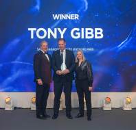 Three people stand on stage, one holding an award. The screen behind them reads "Winner Tony Gibb.