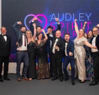 A group of people dressed in formal attire celebrate together at an event, standing in front of a backdrop that reads "Audley Spirit Awards.