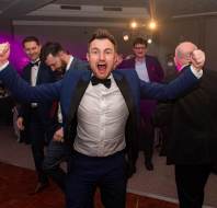 A man in a suit celebrates with arms raised at a formal event, surrounded by other people in suits.