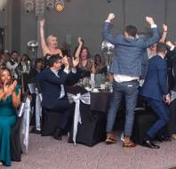 People celebrating and clapping around a table at an event, with some individuals standing and raising their arms in excitement.
