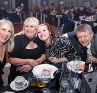 Four people smiling at a formal event, sitting around a table with drinks and plates in a dimly lit room.